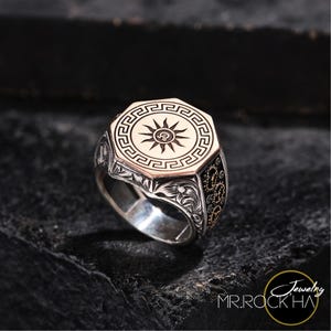 Puede incluir: Un anillo de plata con una parte superior octogonal dorada que presenta un diseño de sol con un borde de clave griega. El anillo tiene intrincados grabados florales en la banda.