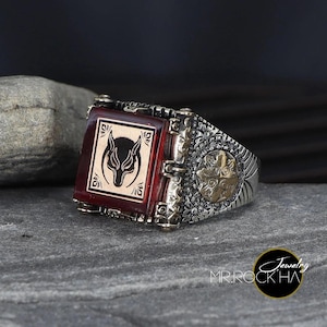 Puede incluir: Un anillo de plata con una piedra roja que presenta un diseño de lobo. El anillo tiene detalles intrincados y un acento dorado.