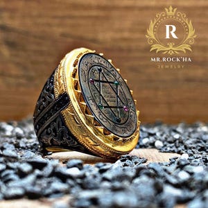 May include: A gold ring with a black and silver engraved band. The ring features a large oval-shaped top with a silver star of David surrounded by Arabic script and five gemstones.