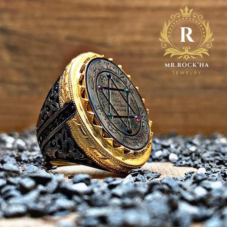 May include: A gold ring with a black and silver engraved band. The ring features a large oval-shaped top with a silver star of David surrounded by Arabic script and five gemstones.