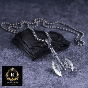 Puede incluir: Un collar de plata con un colgante de hacha detallado. El hacha tiene una superficie texturizada y una cadena de cuentas.