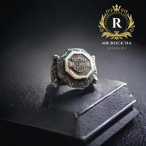 Puede incluir: Un anillo de plata con un diseño detallado que presenta el logotipo de Harley-Davidson en negro y oro. El anillo se muestra en una caja de terciopelo negro.