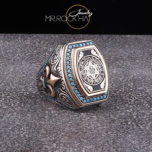 May include: A silver ring with a black and white enamel inlay featuring a Star of David and Arabic script. The ring is surrounded by a row of turquoise blue stones.