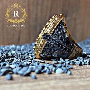 May include: A silver ring with black stones and gold accents. The ring has an intricate design and is set on a bed of small black stones.