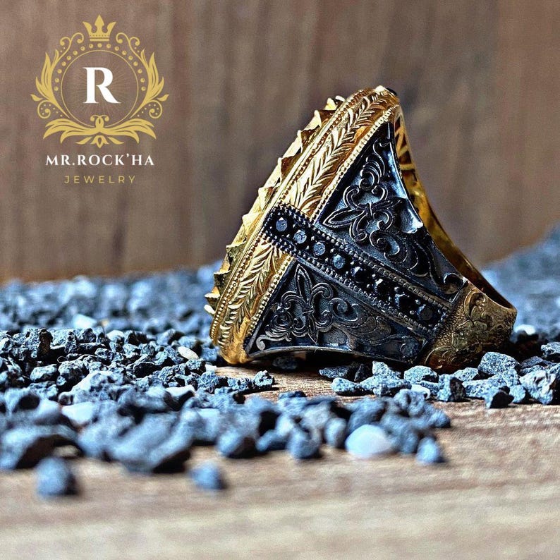 May include: A silver ring with black stones and gold accents. The ring has an intricate design and is set on a bed of small black stones.