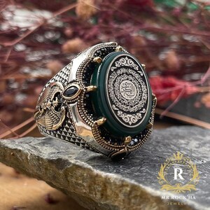 May include: A detailed silver ring with gold accents and a large oval green stone. The stone features intricate black and white calligraphy. The ring has a detailed design on the band and is set on a stone surface.