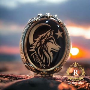 Puede incluir: Anillo dorado con un centro negro ovalado con un diseño de cabeza de lobo, luna creciente y estrella. El anillo tiene un borde ornamentado y detallado y las palabras "MR. ROOKHA JEWELRY" son visibles.