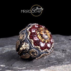 Può includere: Un anello ornato con un motivo a testa di leone. L'anello ha una base in metallo scuro con dettagli dorati e una pietra centrale circondata da gemme rosse e trasparenti. L'anello è posto su una superficie di pietra grigia, con il testo "MR.ROCKHA Jewelry".
