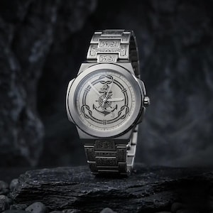 May include: A silver wristwatch featuring an anchor design on the watch face and a patterned strap. The watch has a circular face with a detailed nautical emblem. The watch is displayed on a dark, textured surface.