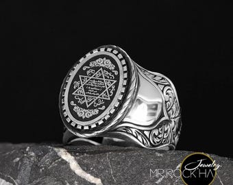 May include: A silver ring with a black oval-shaped center featuring a star of David and Arabic script. The ring band is intricately engraved with a floral design.