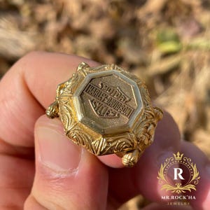 Puede incluir: Un anillo dorado con el logotipo de Harley-Davidson en forma rectangular. El anillo tiene un diseño ornamentado y detallado con una superficie texturizada. El anillo está sostenido en una mano.