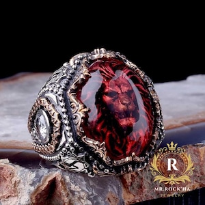 May include: A detailed silver ring with a large, oval, red gemstone featuring a lion's face. The ring has intricate metalwork and a smaller, clear stone on the side. The ring is set on a textured surface.