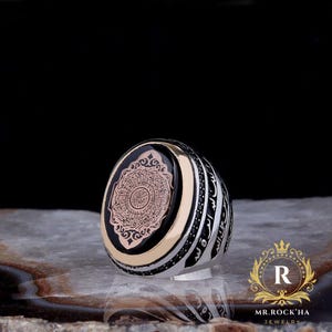 May include: A silver ring with an oval black face featuring intricate gold-colored calligraphy. The ring has a gold band and is accented with black stones. The side of the ring has more calligraphy. The logo "MR. ROCK'HA JEWELRY" is in the bottom right corner.