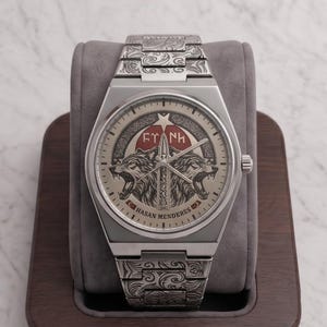 May include: Silver-toned wristwatch with intricate engraved patterns on the band and case. The watch face features a detailed design with two wolf heads, a star, and the text "EY NH" and "HASAN MENDERES". The watch is displayed on a wooden stand.