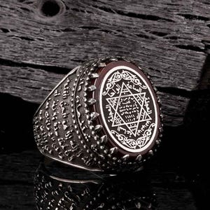 Può includere: Un anello d'argento con una pietra ovale marrone-rossastra. La pietra presenta un design bianco intricato con una stella a sei punte e testo. La fascia dell'anello ha un motivo strutturato e dettagliato. L'anello è esposto su una superficie scura e strutturata.