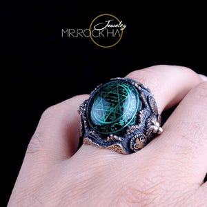 May include: A silver ring featuring a large green gemstone with a pentagram design. The ring is detailed with intricate carvings and a textured finish.