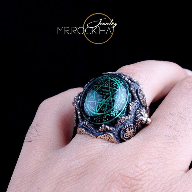 May include: A silver ring featuring a large green gemstone with a pentagram design. The ring is detailed with intricate carvings and a textured finish.