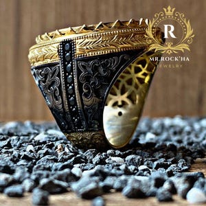 May include: A gold and black ring with intricate designs and black gemstones. The ring is set against a background of small black and white stones.