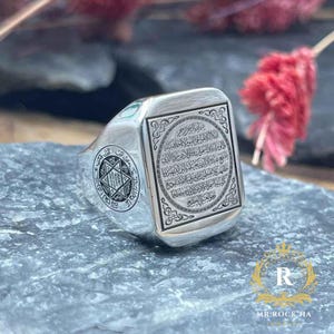 May include: A silver ring with an engraved rectangular face featuring Arabic script and ornate borders. The ring has a square face and a circular design on the side with a star. The ring is displayed on a gray stone surface.
