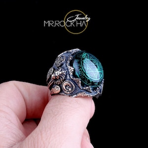 May include: A silver ring with a green stone featuring a pentagram design. The ring has intricate details including a lion head and other decorative elements.