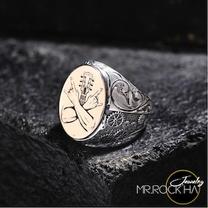 Puede incluir: Un anillo de plata con un diseño ovalado que presenta una guitarra y dos manos haciendo el gesto de rock and roll. El anillo tiene detalles intrincados y un acabado texturizado.