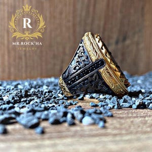 May include: A gold and black ring with intricate designs and black gemstones. The ring is lying on a bed of small black stones.