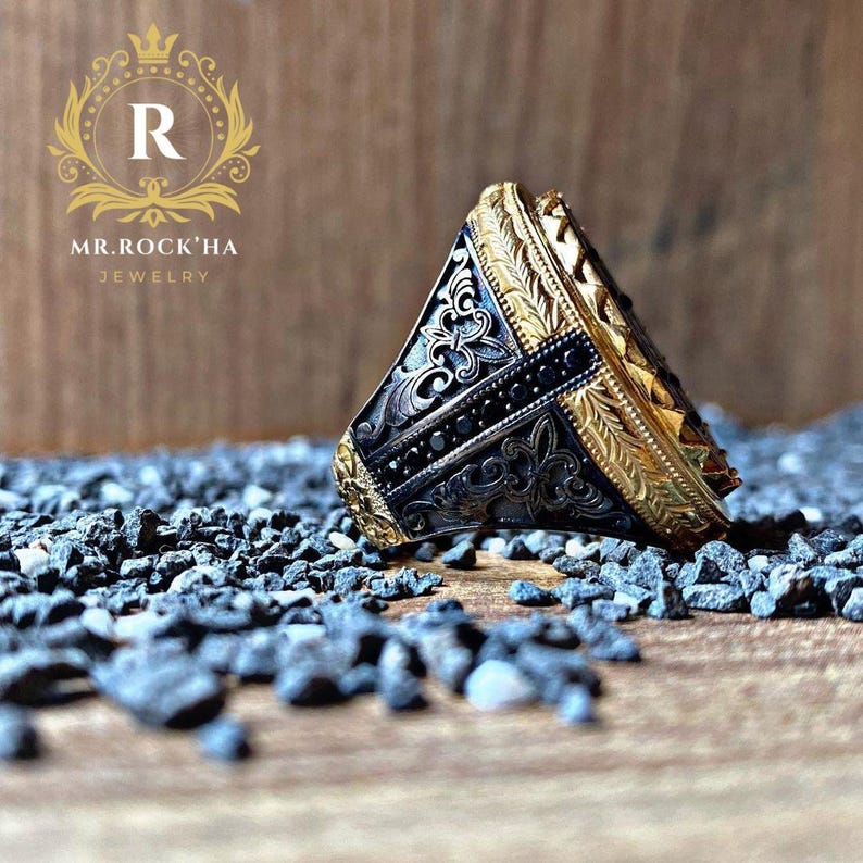 May include: A gold and black ring with intricate designs and black gemstones. The ring is lying on a bed of small black stones.