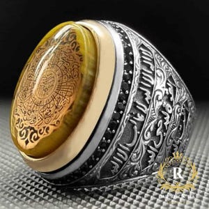 May include: A detailed silver ring with a large, oval, yellow-brown stone. The stone features intricate gold-colored calligraphy. The ring has a gold band around the stone and is accented with black stones and ornate silver detailing.