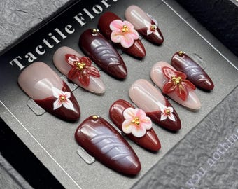 Può includere: Un set di unghie finte con una varietà di design. Alcune unghie sono di un bordeaux intenso, altre sono nude con una punta bordeaux. Diverse unghie presentano decorazioni floreali 3D con accenti dorati. Il testo "Tactile Flora" è visibile.