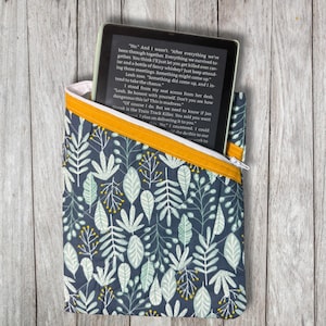 May include: A quilted e-reader sleeve with a navy blue floral pattern and a yellow zipper. The sleeve holds an e-reader displaying text. The floral design includes white, light green, and yellow accents. The sleeve is on a wooden surface.
