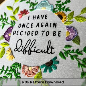 May include: Embroidered hoop art with a white background and green floral embroidery. The text "I have once again decided to be difficult" is embroidered in black thread.
