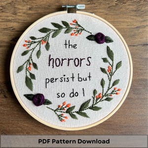 May include: Embroidered hoop art with a floral wreath and the text "the horrors persist but so do I".