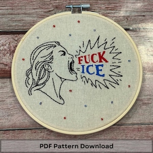 May include: Embroidered hoop art featuring a woman's profile with an open mouth, screaming the words "FUCK ICE" in red and blue. The design is on a beige fabric, accented with red and blue beads, and set in a wooden hoop.