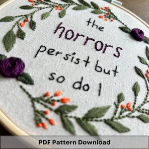 May include: Embroidered hoop art with a floral wreath and the text "the horrors persist but so do I".