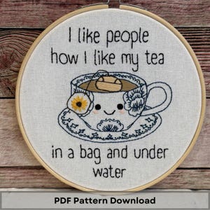 May include: Embroidered hoop art with the text "I like people how I like my tea in a bag and under water." The design features a smiling teacup with a yellow flower and blue floral details. The hoop is set in a wooden frame.
