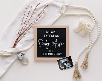 May include: A pregnancy announcement with a black letter board that reads "WE ARE EXPECTING Baby Aspen DUE DECEMBER 2022". The image includes a sonogram, baby booties, a wooden bead garland, a crochet rattle, and decorative branches.