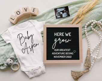 Puede incluir: Un body blanco con el texto "Baby Aspen" en escritura cursiva negra. Un tablero de letras con el texto "Here we grow. Our greatest adventure begins November 2022" está al lado del body. También hay una guirnalda de cuentas blancas con borlas, un pequeño elefante de peluche y un bloque de madera que deletrea "LOVE" en la imagen.