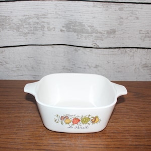May include: White square baking dish with a floral design and the text "Le Creuset" on the bottom. The dish has two handles on the sides.