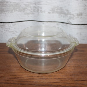 Puede incluir: Un plato para hornear de vidrio transparente con tapa. El plato es redondo y tiene dos asas a los lados. La tapa es ligeramente abovedada.