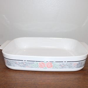 May include: White rectangular baking dish with handles. The dish has a decorative band with a floral pattern of pink roses and green leaves. The dish is sitting on a brown wooden surface.