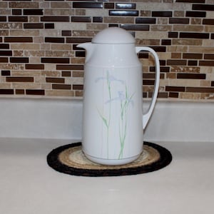 May include: A white thermal carafe with a rounded lid, a curved handle, and a spout. The body of the carafe features a delicate, light-colored botanical illustration of thin, tall stems with pale blue flowers extending vertically along the front.