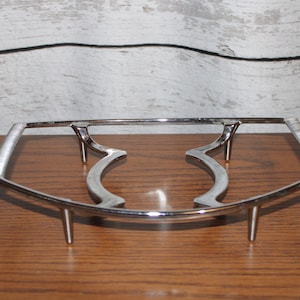 May include: A silver metal hot plate stand with a curved design and three legs.