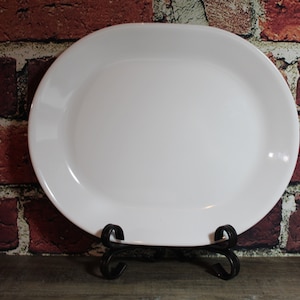 Puede incluir: Una fuente ovalada grande de cerámica blanca. El plato se exhibe sobre un soporte decorativo de metal negro. El fondo presenta una pared de ladrillos.