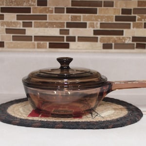 May include: A brown glass saucepan with a lid and a handle, resting on a round, woven trivet. The cookware is transparent, allowing a view of the interior. The background features a decorative tile backsplash.