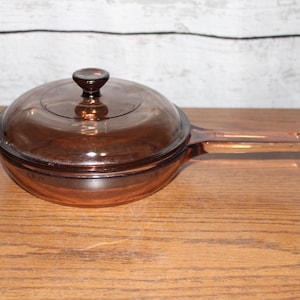 May include: A vintage amber-colored glass saucepan with a matching lid. The pan has a long handle and a round shape. The lid has a small knob. The cookware is made of transparent glass.