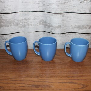 Puede incluir: Tres tazas de cerámica azules con asas redondeadas. Las tazas están dispuestas en fila sobre una superficie de madera.
