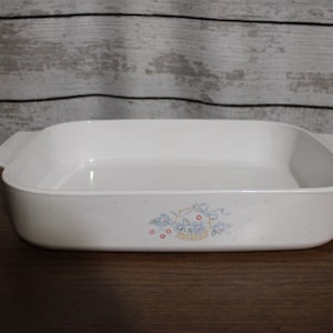 May include: White ceramic baking dish with a floral design featuring blue flowers and a basket. The dish has a square shape with rounded corners and two handles.