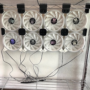 May include: Eight white computer fans with black accents are mounted on a metal shelf. The fans are connected to wires and are arranged in two rows of four.