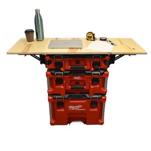 May include: A red and black Milwaukee Packout toolbox with a wooden work surface attached. The work surface is supported by a metal frame and folds down. The toolbox is stacked on top of two other Milwaukee Packout toolboxes.