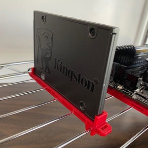 May include: A black Kingston solid state drive (SSD) with a red mounting bracket. The SSD is mounted to a metal shelf.
